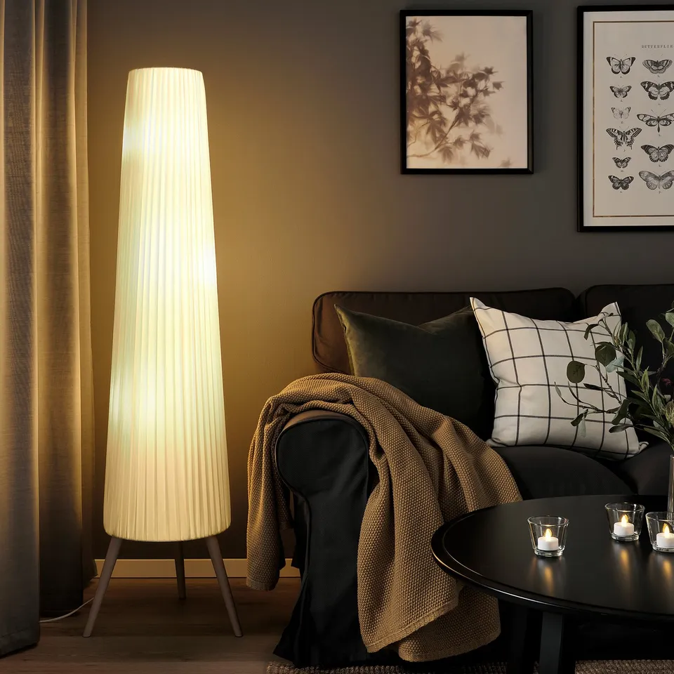 Cosy living room with tall white ÖKENSAND lamp, dark sofa, warm cushions & candles. Artwork on walls, light brown curtains.