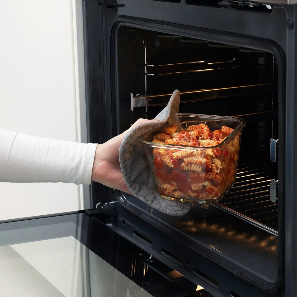 Person placing pasta dish into oven, showcasing oven-safe glass and heat-resistant lid.