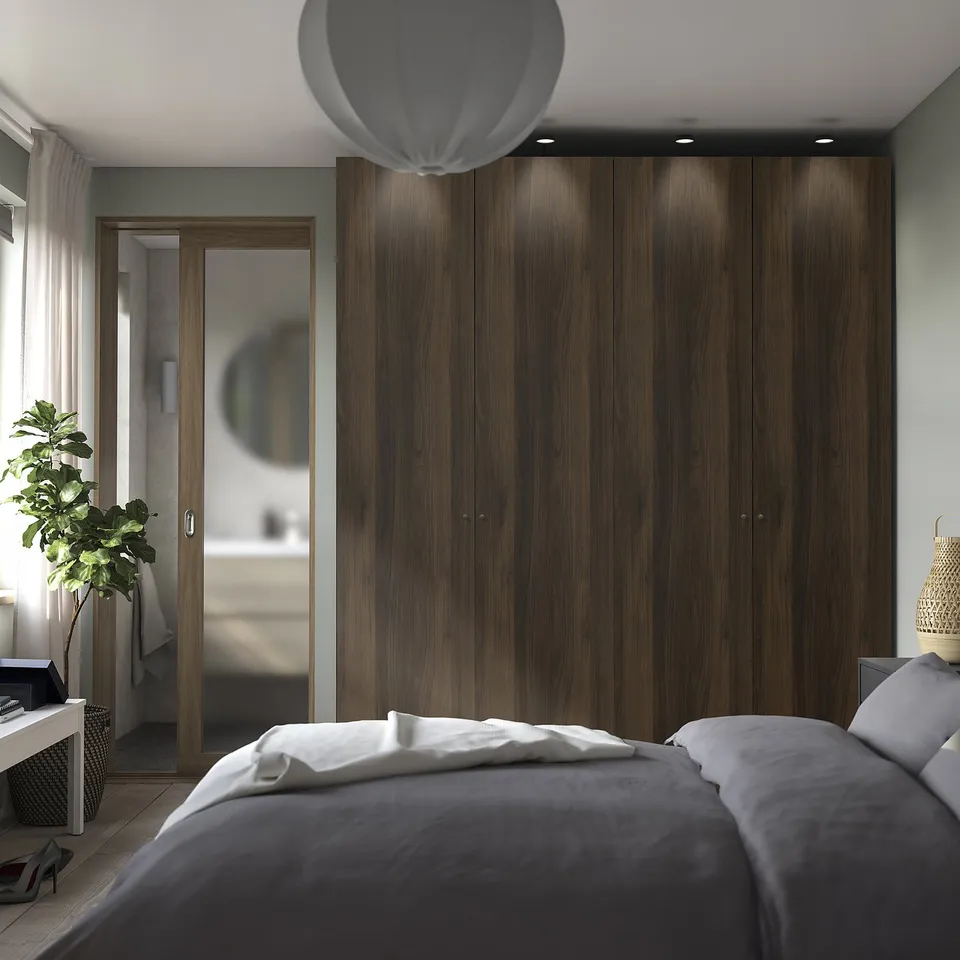 Modern bedroom with large dark wood PAX wardrobe. Bed has grey bedding, beside plant and lamp. Wardrobe towers over room, reflecting light.