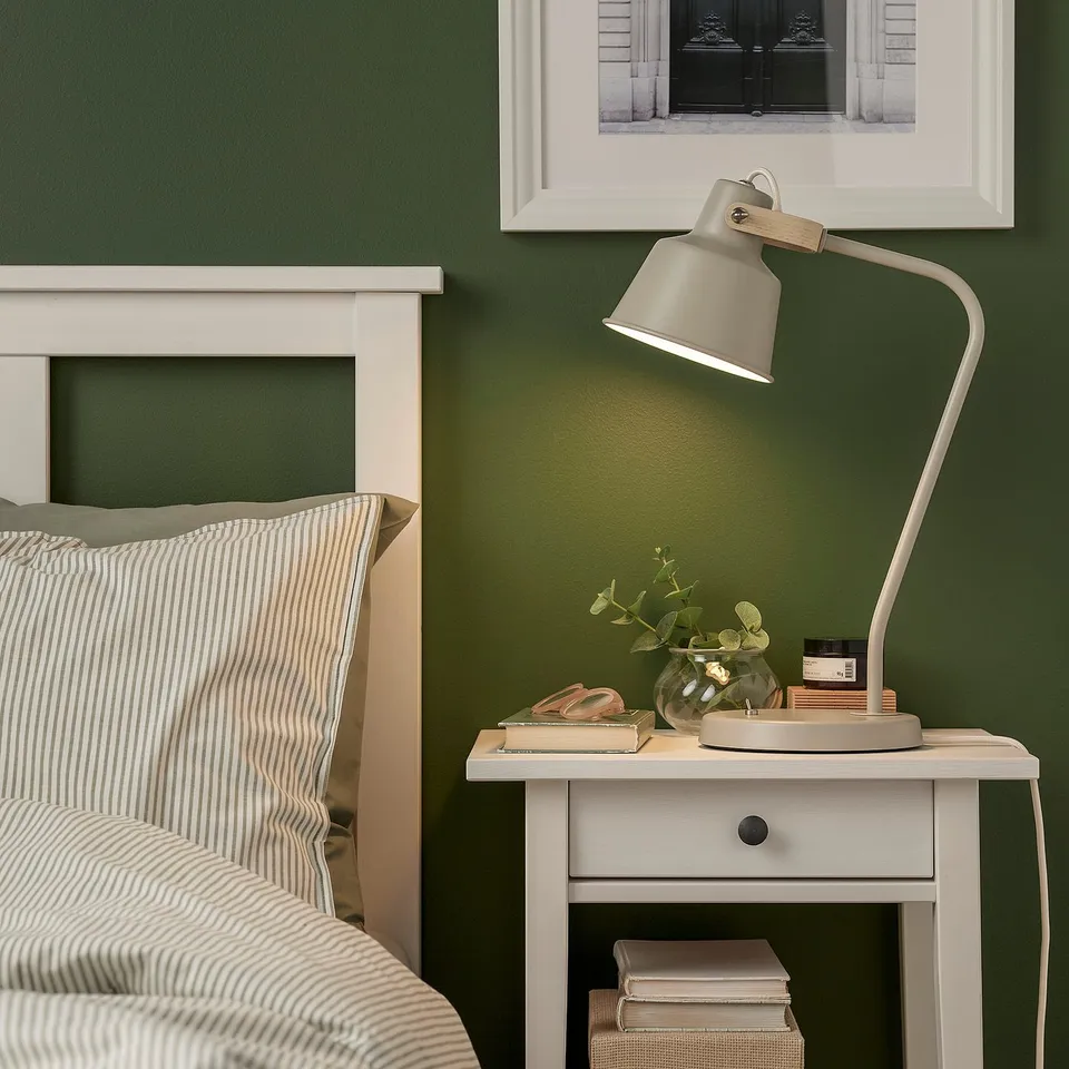 SJÖMAKT desk lamp on bedside table with books, vase, and striped pillows. Dark green wall.