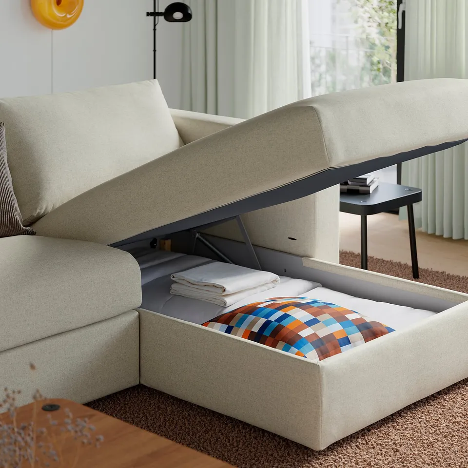 Beige sofa with open storage compartment showing folded blanket and pillows, minimalist room.