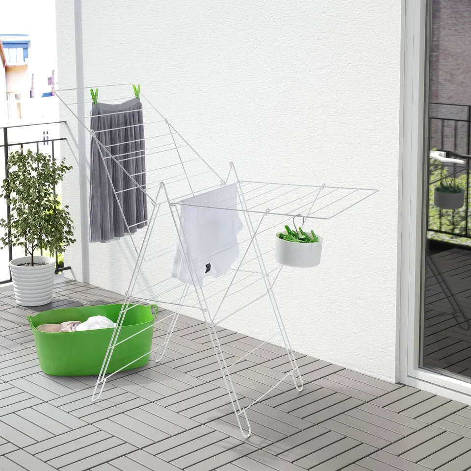 White FROST drying rack on balcony, larger than nearby laundry basket, holds clothes & hanging plant.
