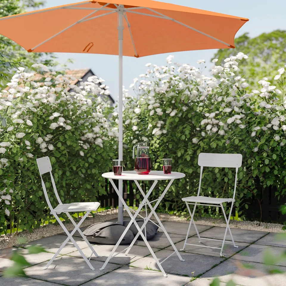 A garden scene with a white table and chairs under an orange HÖGÖN parasol, surrounded by blooming bushes.