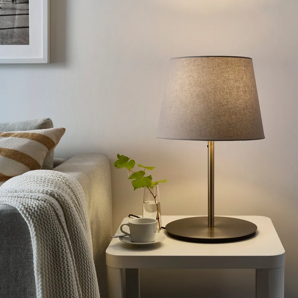 SKOTTORP beige lamp on side table with vase and cup. Couch with blanket and pillow in background.