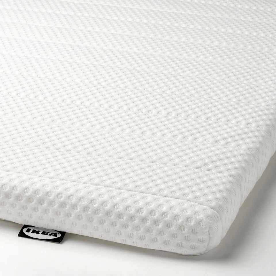 Closed foam mattress topper with a textured, white surface and a small tag labelled IKEA on the edge.