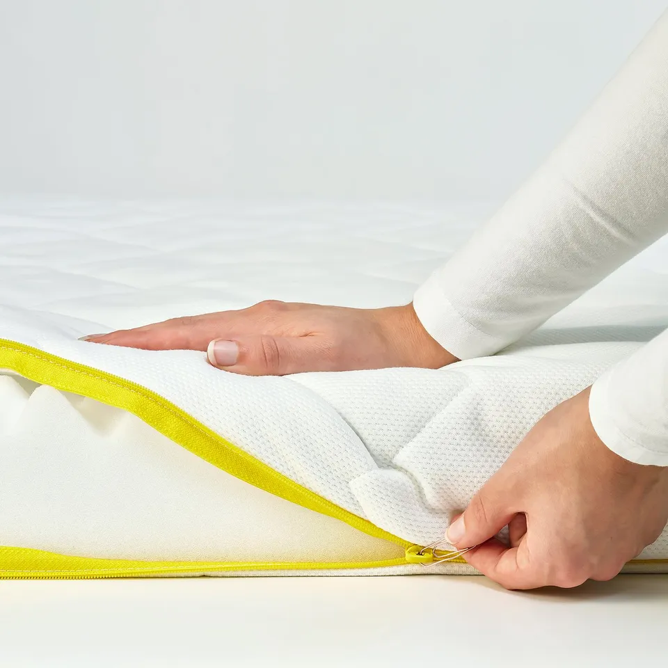 Hands opening a yellow zip on a white mattress cover, highlighting its child-safe design.