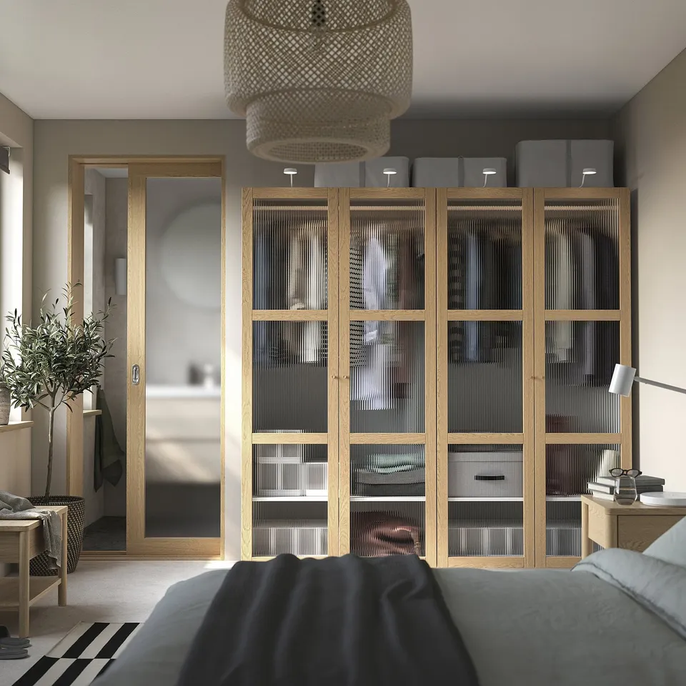 Large bedroom with wooden cabinet, glass doors, mirror, bed, bedside table, lamp, plant, and wicker light fixture.