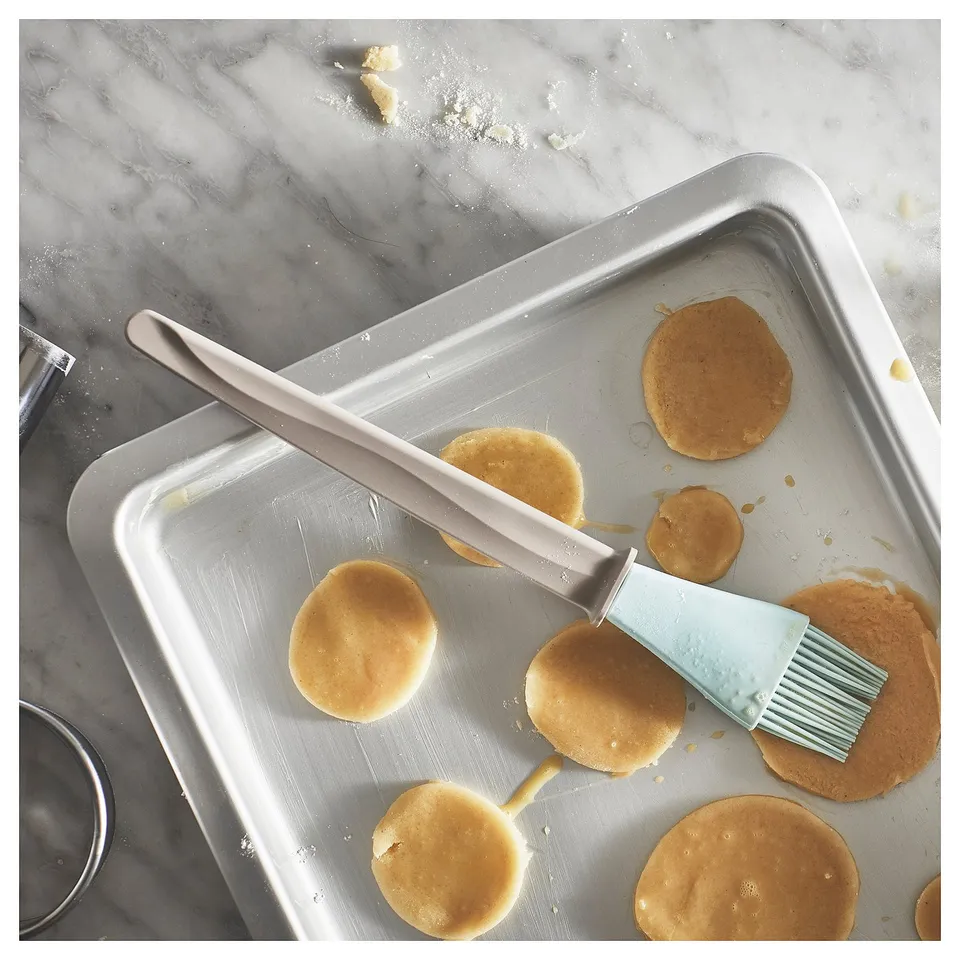 Silver baking brush resting on tray with pancakes. Long handle, blue bristles, used for applying oils.
