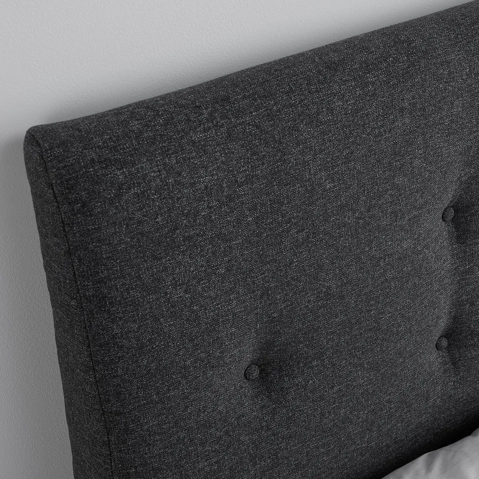 Its a close-up of a dark grey upholstered bed headboard with neat button tufting, showing its high-quality fabric and design.