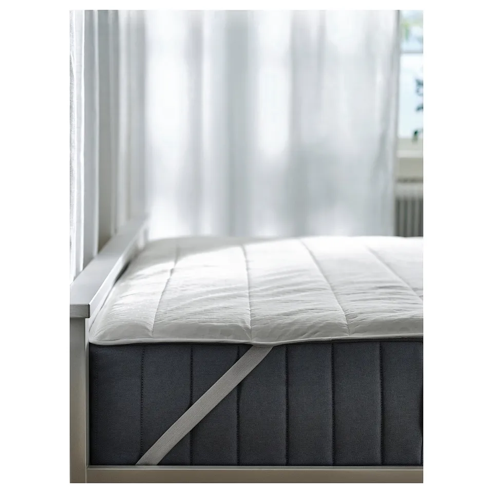 A white ROSENVIAL mattress protector placed on a bed, featuring a soft, quilted design, and a grey strap securing it.