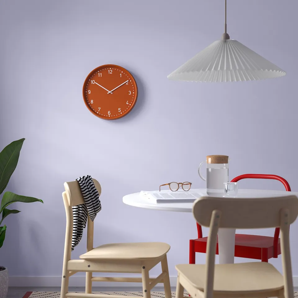 Modern kitchen with orange-silver clock BONDIS on grey wall, round table, red chairs, plant, and pendant lamp.