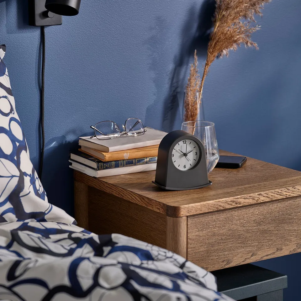 Bedside bedside table with a wooden top holding an alarm clock, books, glasses, a glass with dried flowers, and a phone against a blue wall.