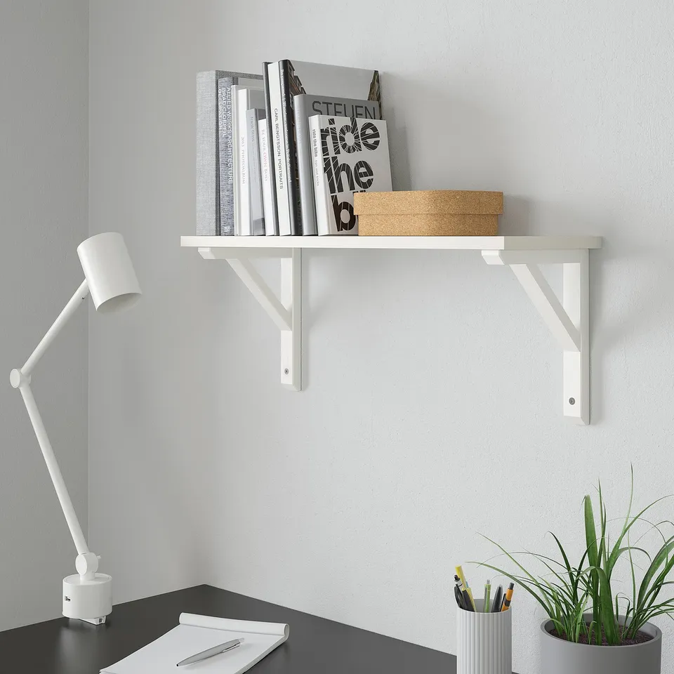 White BURHULT shelf holds books, box, and plant. Mounted high above desk with lamp and papers.
