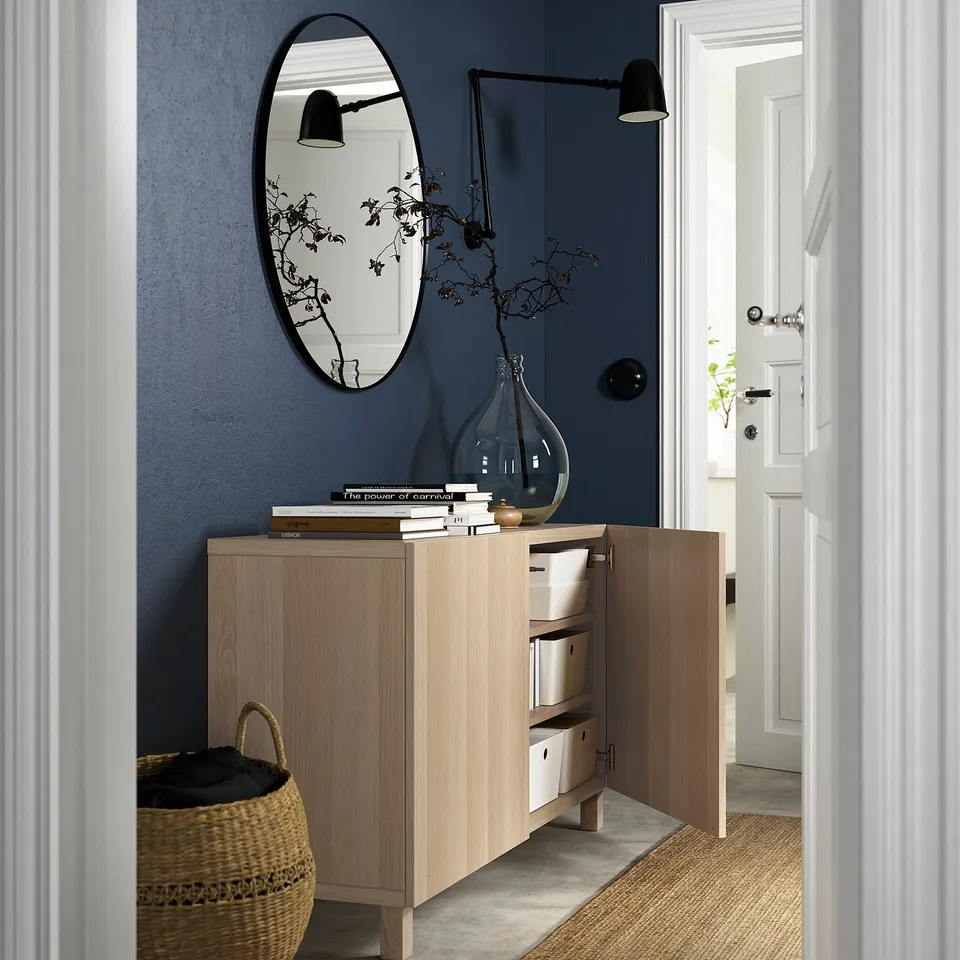 Open light wood sideboard with white boxes. Dark blue wall, round mirror, and wall lamp.