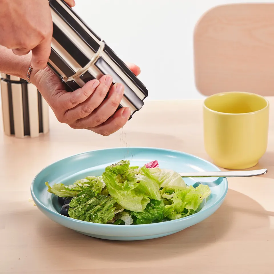 Hand grinding salt over salad. Black and white striped salt mill, blue plate, yellow cup.