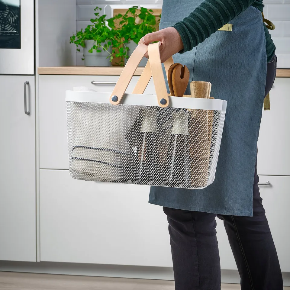Person holding wire basket with wooden handles, containing kitchen utensils, in a kitchen. The baskets mesh design allows contents to be visible.