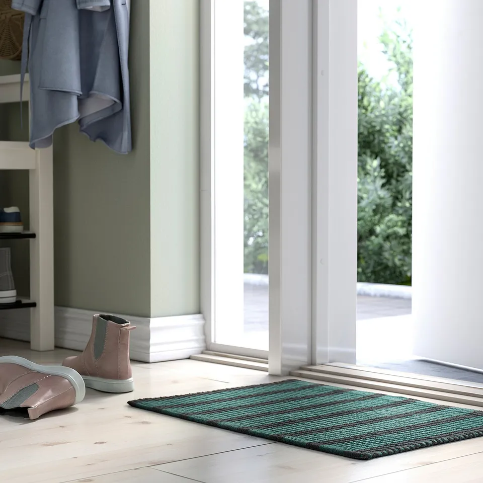 Doormat named VÄGSKYLT in blue-green & black stripes, cotton, rectangular, placed near a door with pink boots and a coat, smaller than the boots.