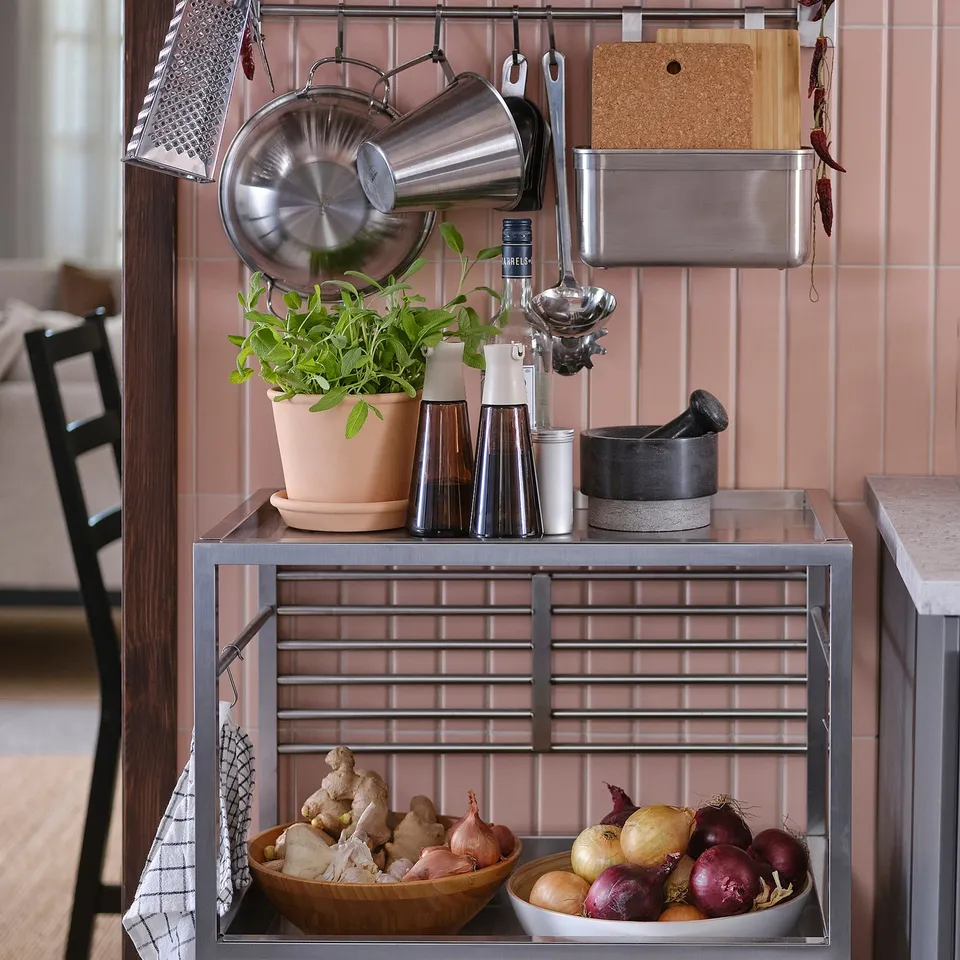 Kitchen cart with shelves: pots, plant, oils, spices & onions. Pink tiled wall.