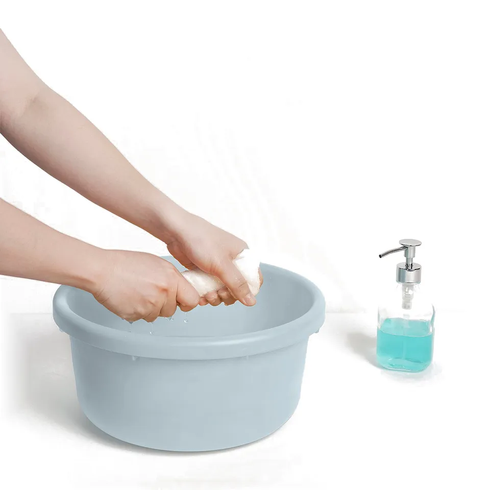 A person washing hands in a blue plastic bucket with soap. A soap dispenser with blue liquid stands nearby.