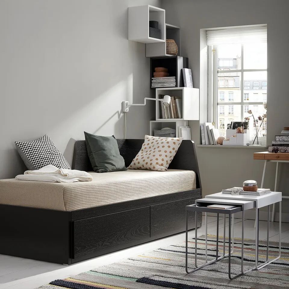 Modern living room with FLEKKE grey sofa-bed, white cushions, black side tables, striped rug, white bookshelf, and large window.