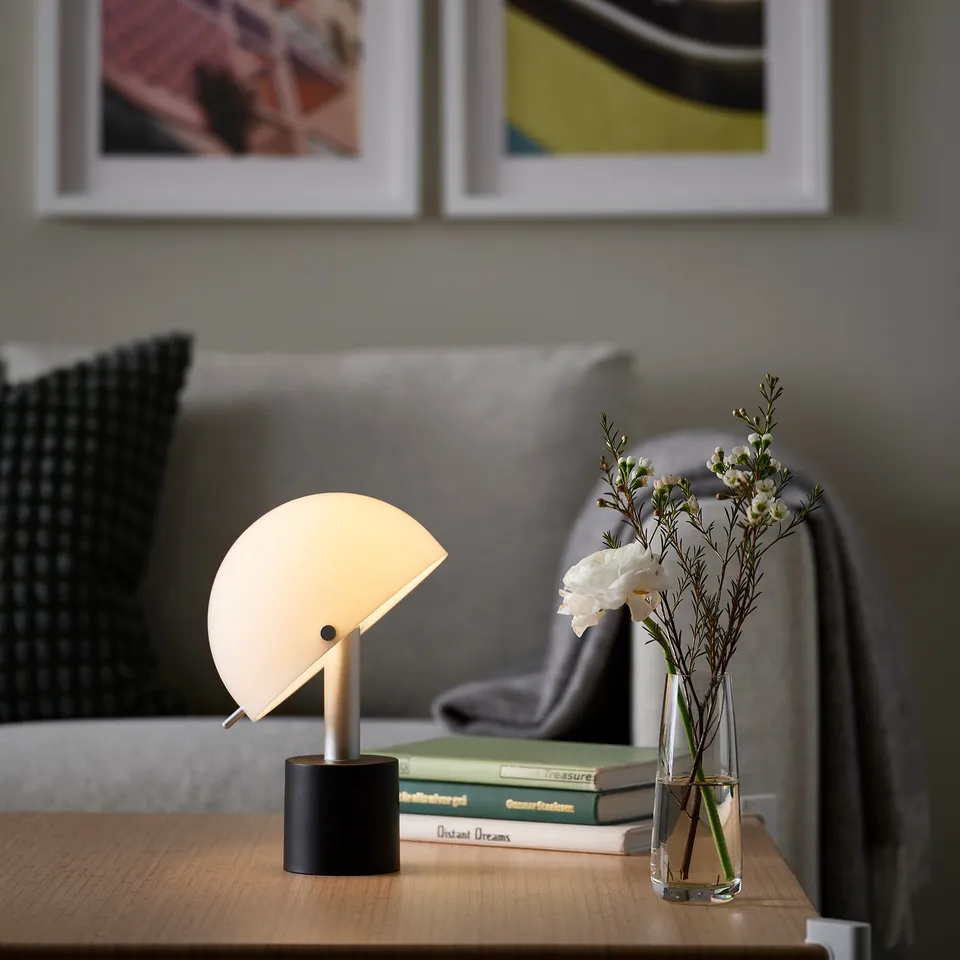 Small NÖDMAST lamp on table beside grey sofa, with books and flowers. Bright light, sleek design.