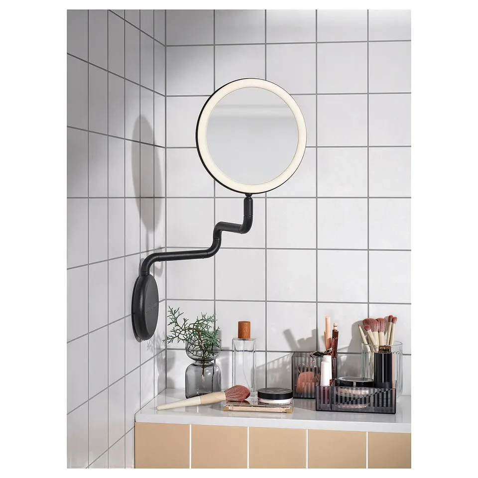 Modern black mirror with built-in lights, mounted on a wall, surrounded by makeup accessories on a white tiled bathroom counter.