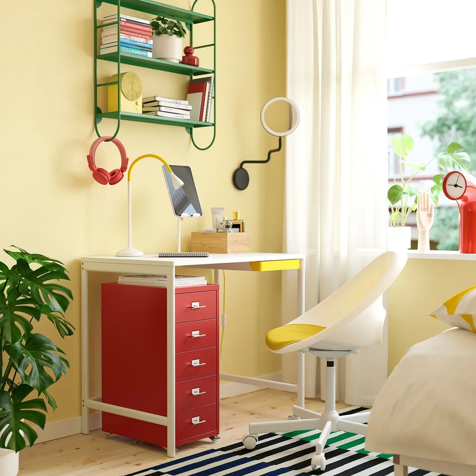 Vibrant home office with yellow walls, featuring ENKROKEN red desk, white chair, green shelves, and plants.