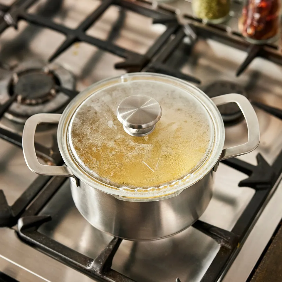 A saucepan on a stove, bubbling with liquid, covered with a glass lid featuring a steam release valve.