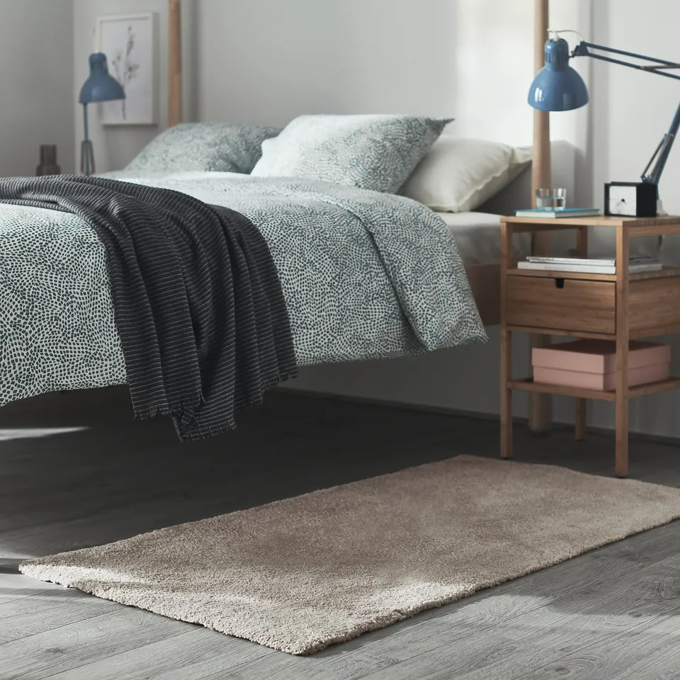 Modern bedroom with grey rug, wooden bedside table, lamp, and bed with patterned pillows and blankets.