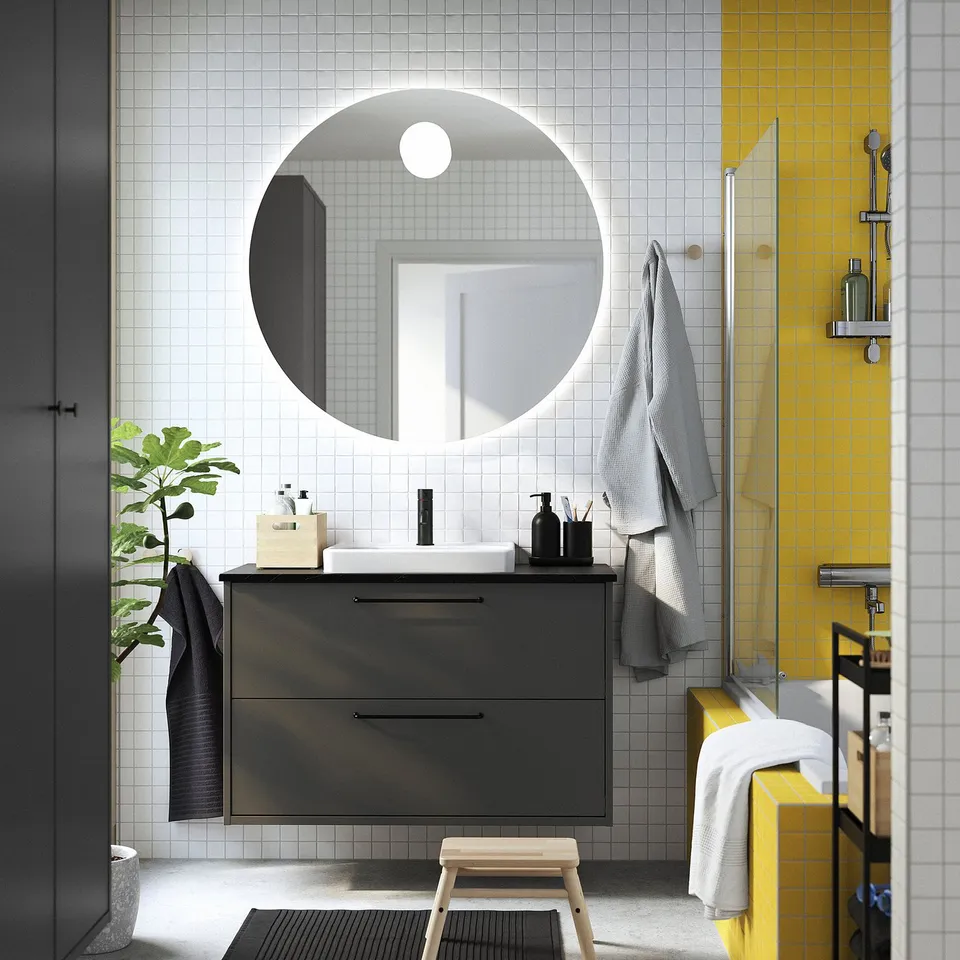 Modern bathroom with dark grey HAVBÄCK vanity. Large round mirror, white sink, yellow tiles, and grey towels. Plant and stool included.