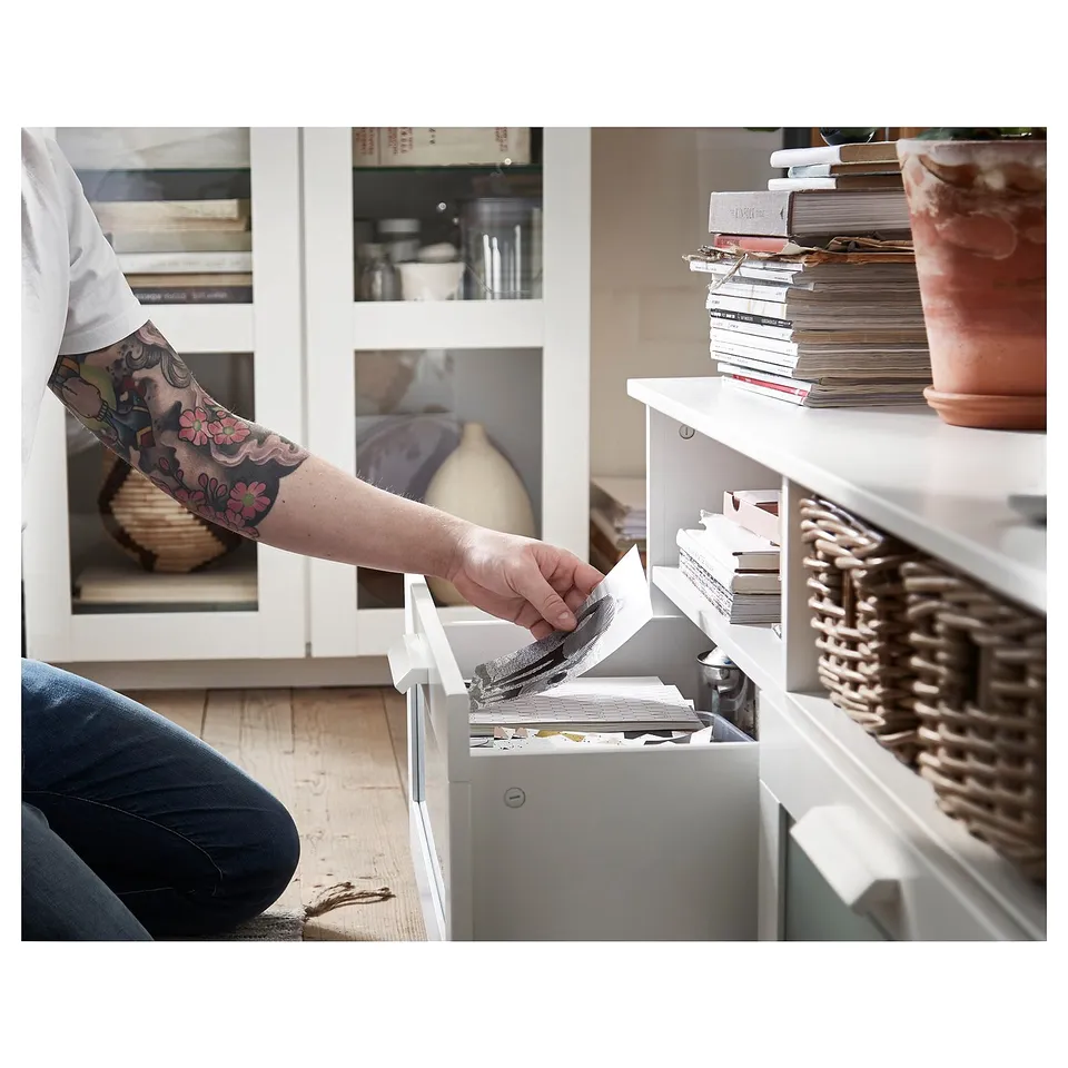 Person with tattooed arm reaches into white tv unit drawer; shelves filled with items, plant, and books.