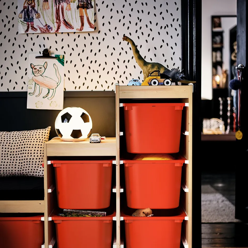Bright red storage bins, wooden frame. Wall: patterned, childs art. Top: football, dinosaur toy. Left: cushions, cat drawing. Right: bookcase, medal.