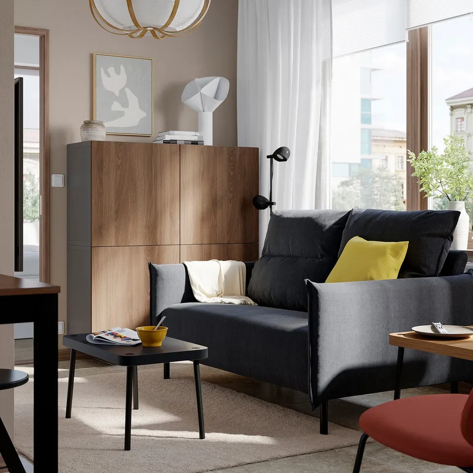 Modern living room with MANNARP dark grey sofa, wooden cabinet, black side table, yellow pillows, and large windows.
