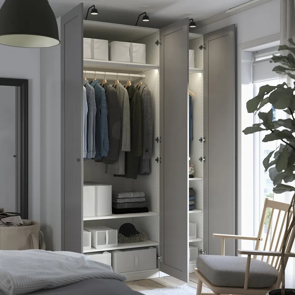 A modern bedroom an open grey PAX wardrobe displaying clothes on hangers, shelves with storage boxes, and integrated lighting.