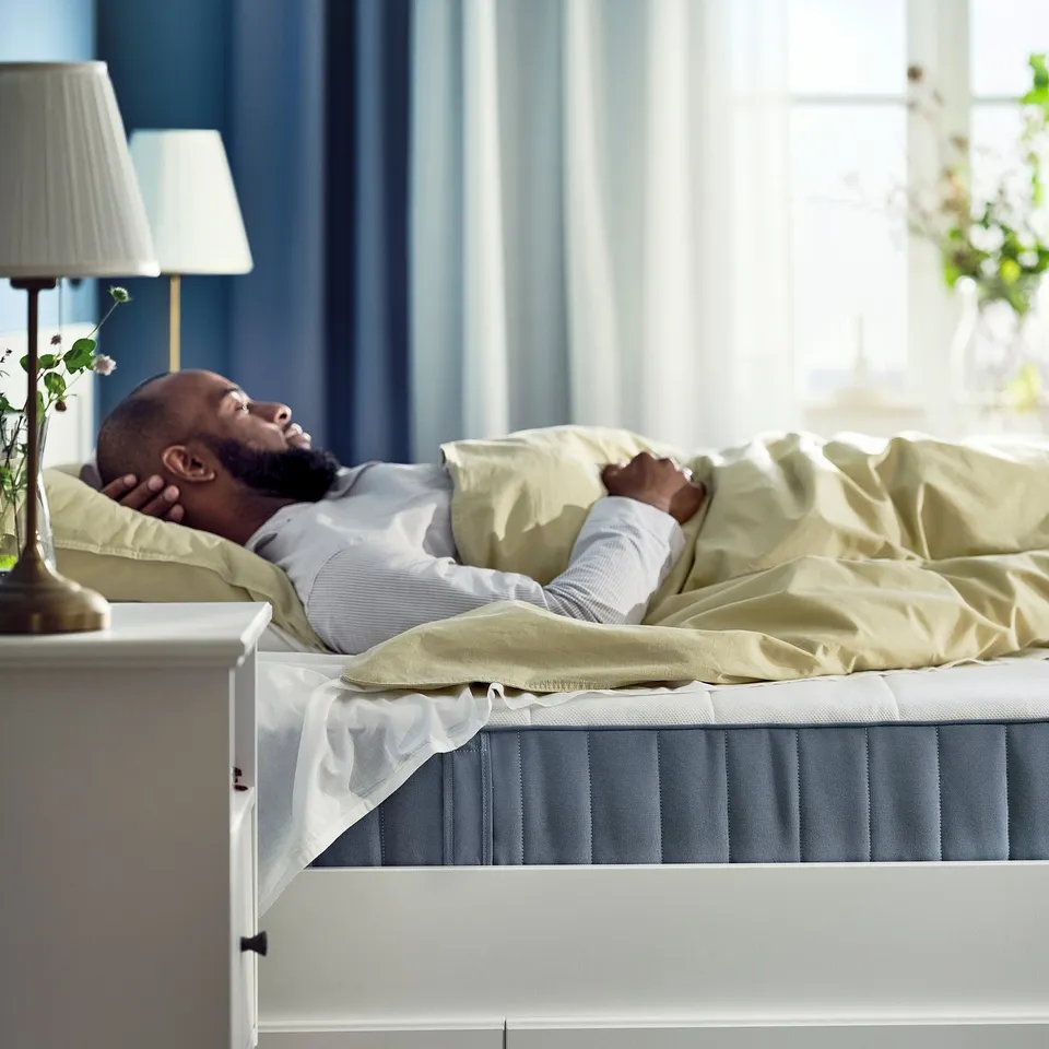 Person sleeping on white VESTERÖY mattress with blue bottom, light yellow pillows, beige blanket, bright natural light.
