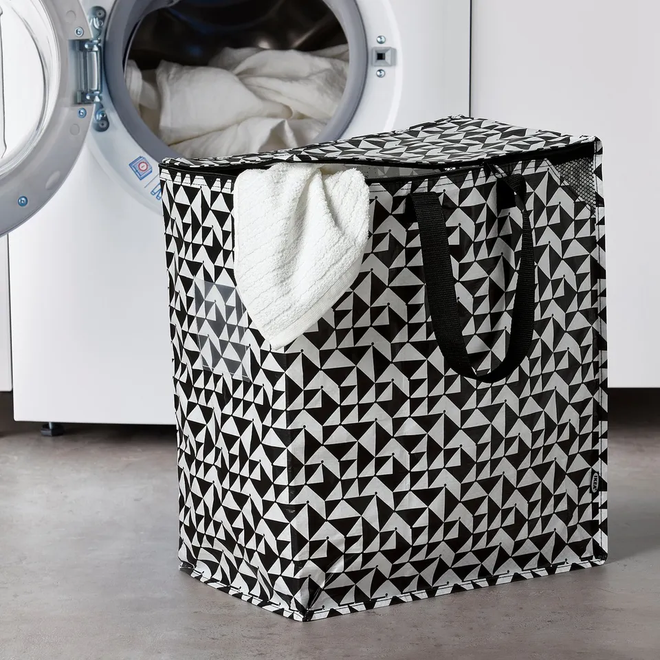 KNALLA laundry bag with geometric pattern next to washing machine. Durable polyester, zips closed, compact yet spacious.