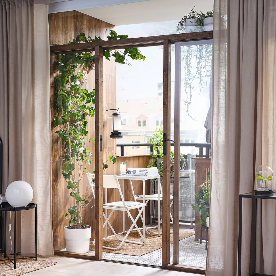Modern bedroom with large window, sheer curtains, potted plants, and outdoor furniture set on balcony.