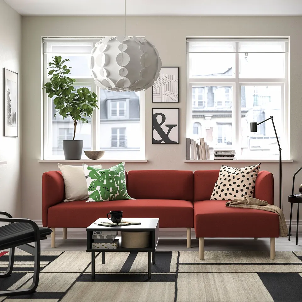 Modern living room with red modular sofa, green cushions, large light, urban view, geometric rug, plant, and wall art.