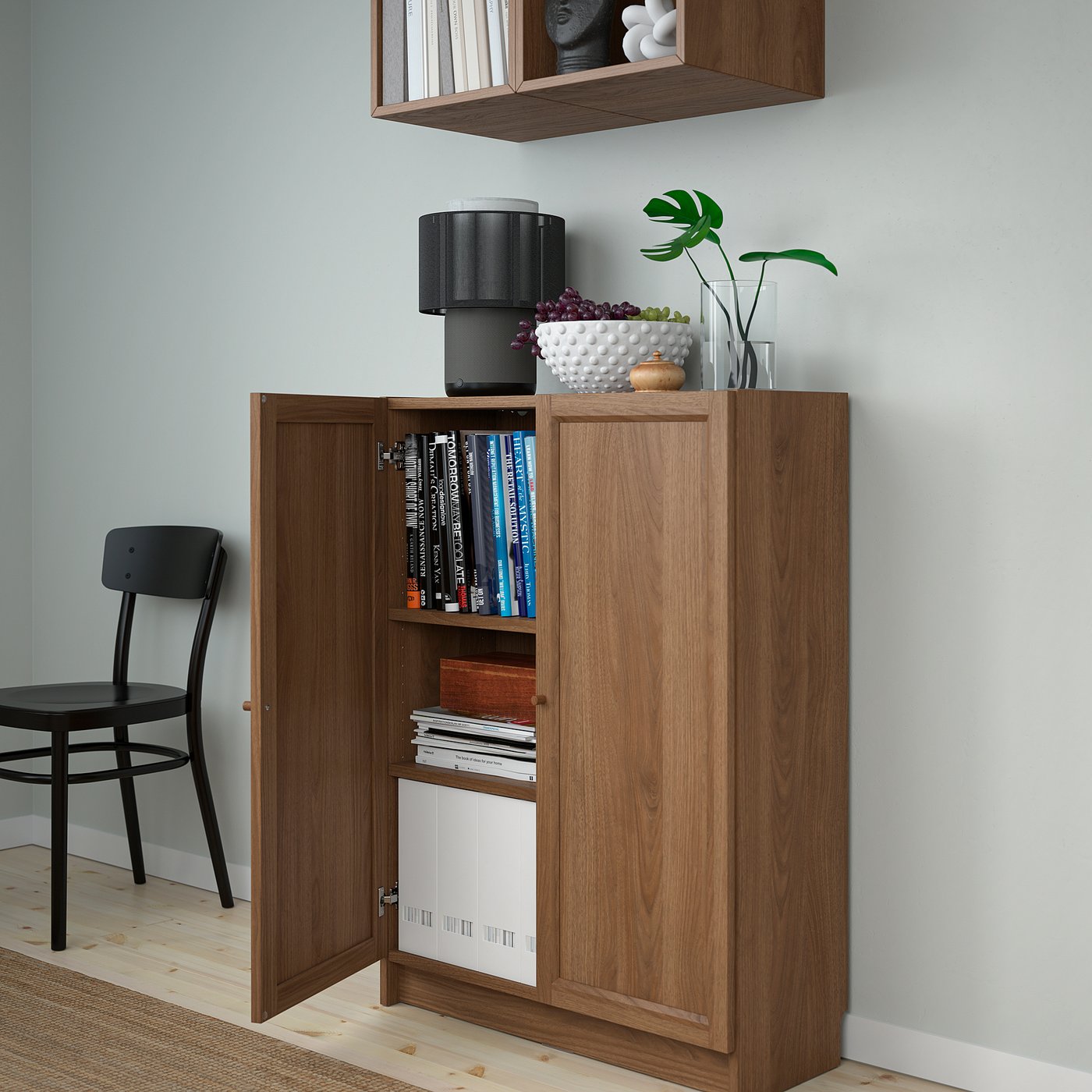 Wooden cabinet with shelves, open doors showing books and magazines, lamp, vase on top, black chair beside.
