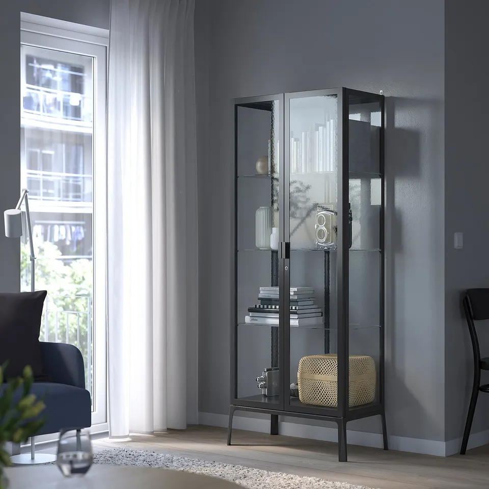 Black MILSBO glass-door cabinet in corner by window with glass shelves and decor.