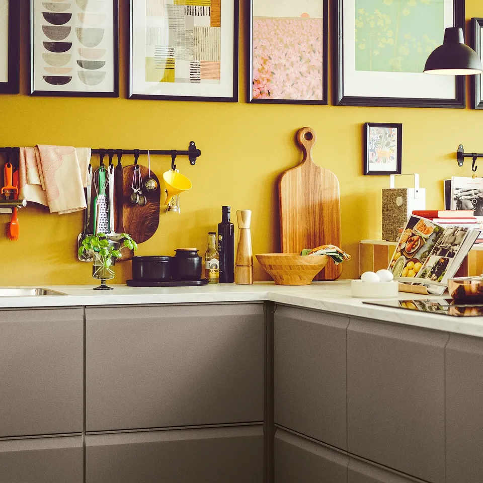 Yellow kitchen with grey cabinets, hanging utensils, plant, colourful art, cookbook, and tall pepper grinder.