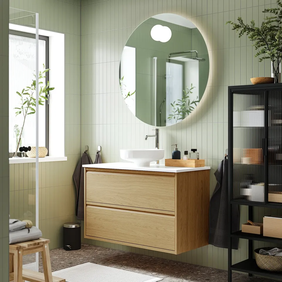 Modern bathroom with ÄNGSJÖN light oak vanity, marble basin, round mirror, window, and shelf. Green walls and dark floor enhance space and warmth.