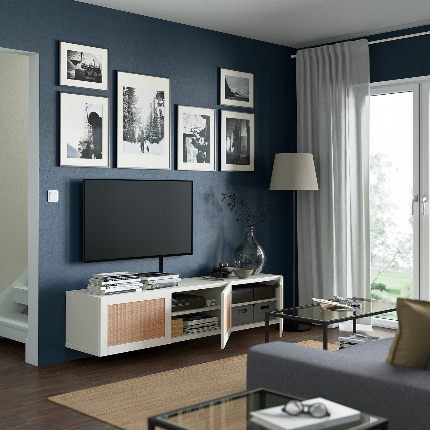 Modern living room: tv on white BESTÅ stand, black framed photos on dark wall, glass coffee table, grey curtains, lamp, and grey sofa.