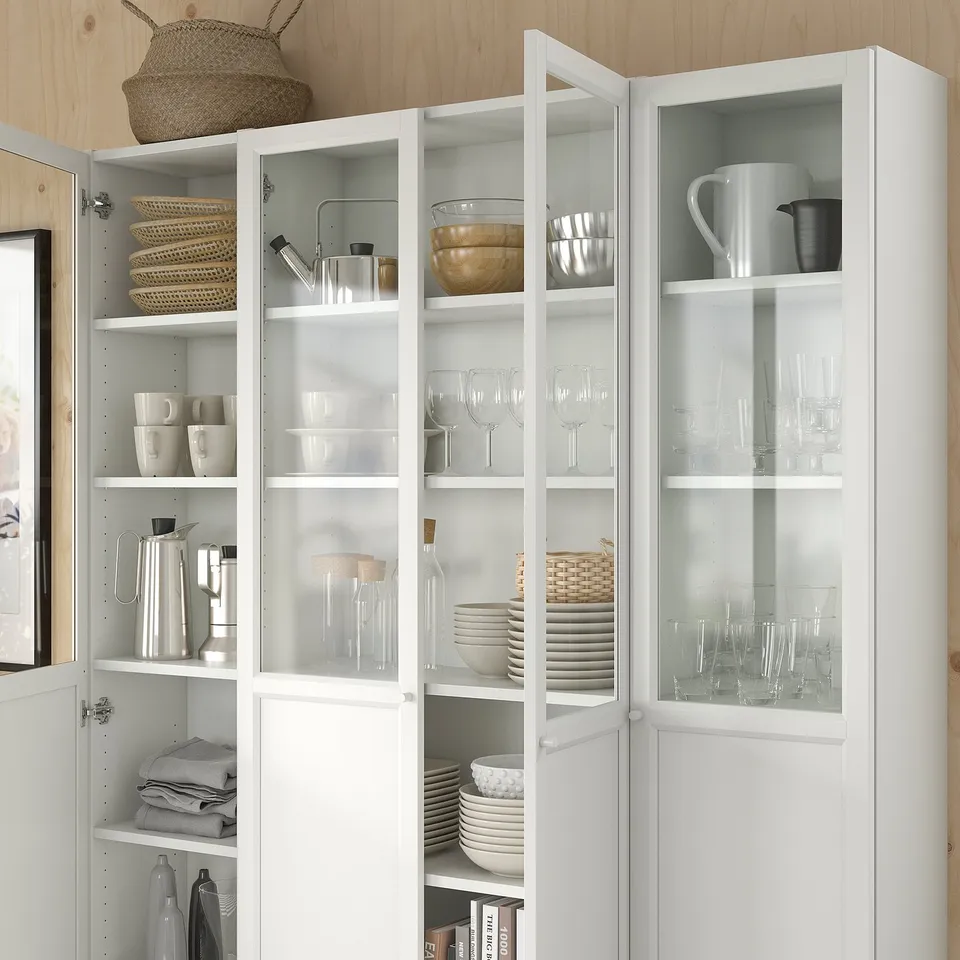 White BILLY bookcase with glass doors; neatly organised kitchen items, dishes, glasses, and linens; minimalist and functional design.