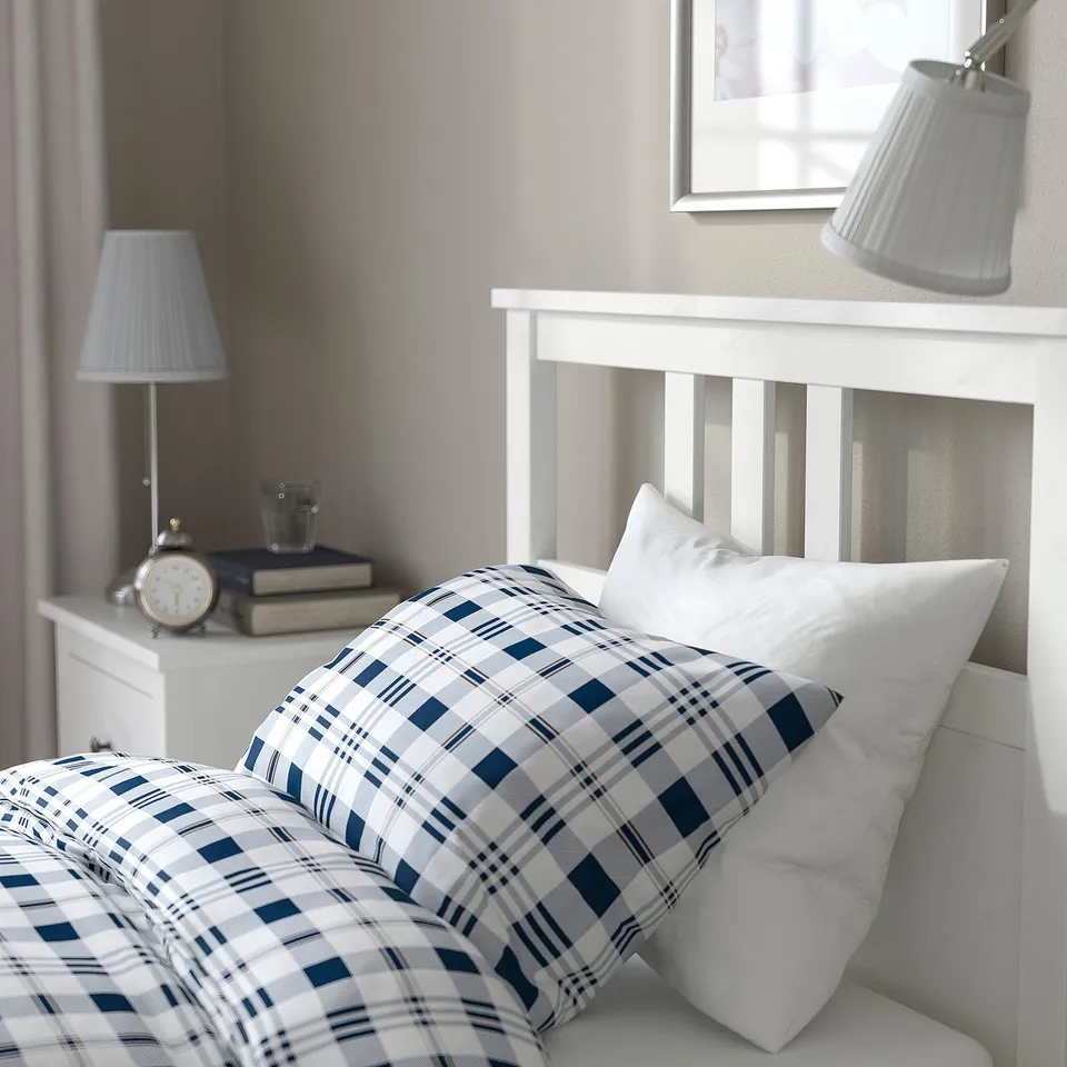 Cosy bed with blue plaid bedding atop a white headboard, accompanied by two nightstands with lamps and a clock, conveying a serene bedroom ambiance.
