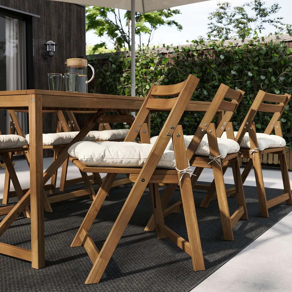 Outdoor wooden patio set with light-colored cushioned chairs neatly arranged on a black rug. The table has a pitcher and cups.