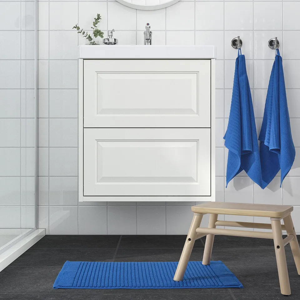 Modern bathroom with white cabinet, glass shower, blue ALSTERN mat & towels on wooden bench.