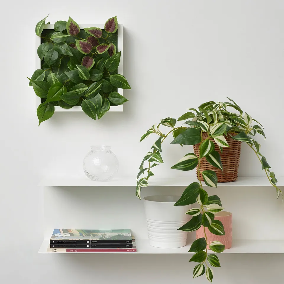 White wall shelf with FEJKA artificial green plant wall panel, basket plant, books, and vases. Panel size similar to books, wall-mounted.