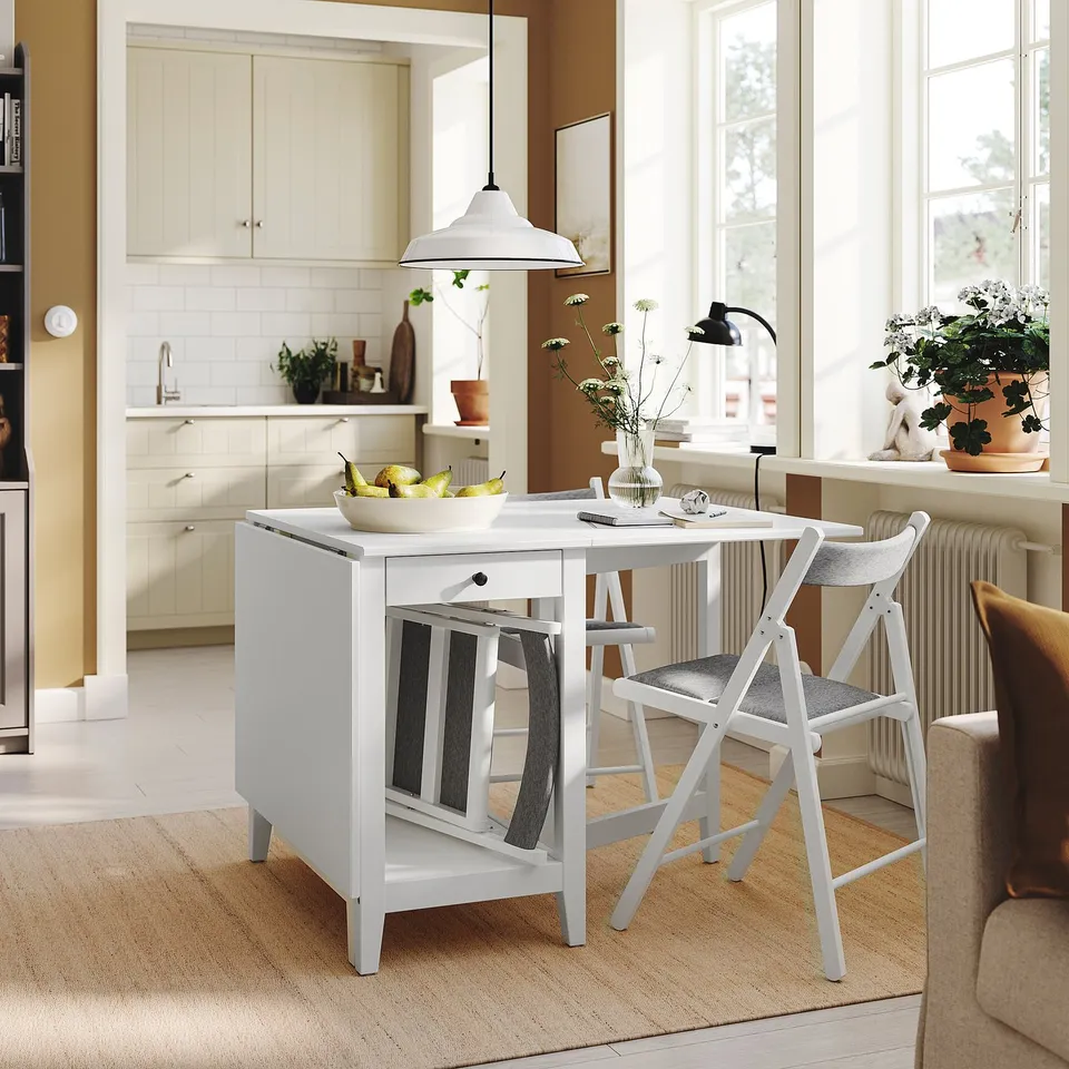 Small white NÄSINGE drop-leaf table with chairs, potted plant, kitchen in background.