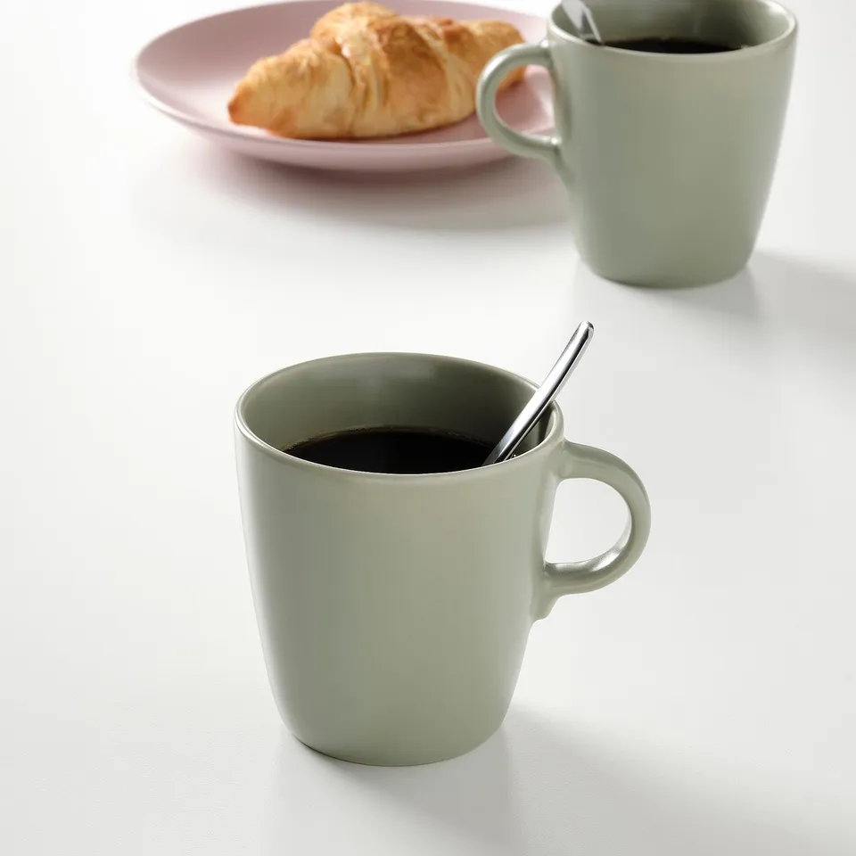 Two mint-green mugs with black coffee, one spoon, and a croissant on pink plate.